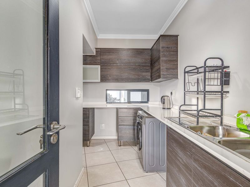 A functional laundry room featuring modern wood-grain cabinetry, stone countertops, a double stainless steel sink, and tiled flooring with dedicated appliance plumbing.