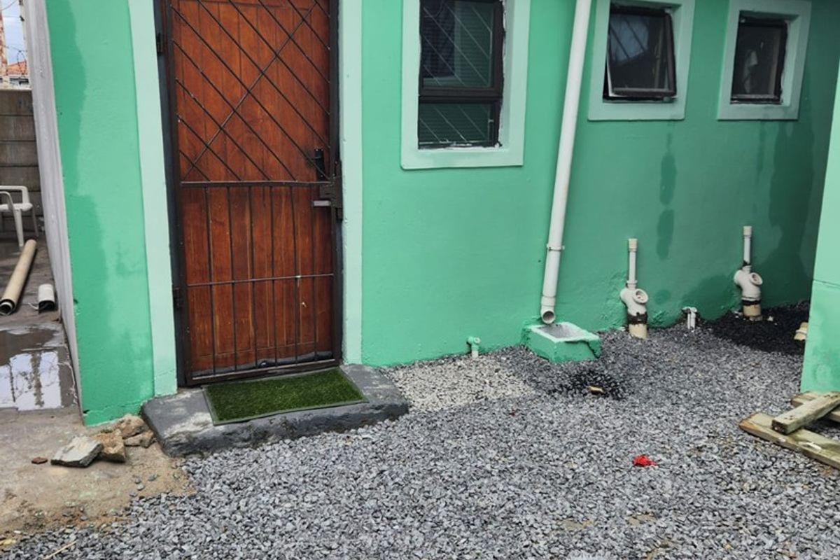 Rear exterior view of the property featuring green painted walls, security gate, and exposed plumbing infrastructure with gravel ground cover.