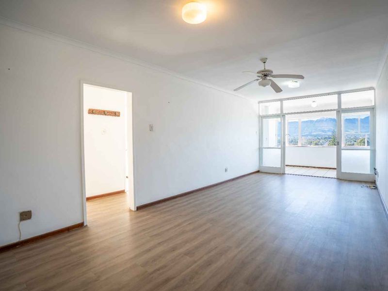 A bright, empty living room featuring laminate flooring, a ceiling fan, and direct access to a balcony with scenic mountain views.