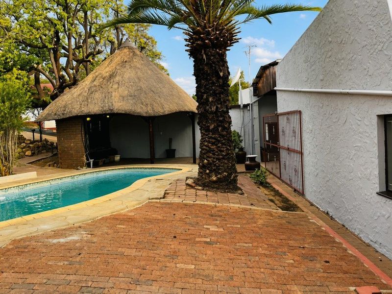 Outdoor pool area featuring a traditional thatch lapa, brick paved patio, and established palm tree, providing a relaxed space for outdoor entertainment.
