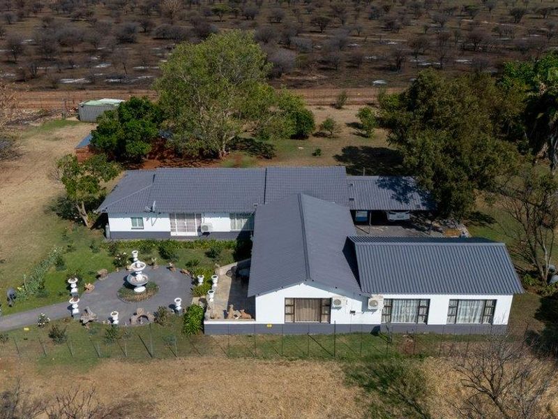 Aerial view of a farm property featuring a main dwelling with a metal roof, paved patio area, and surrounding rural landscape.