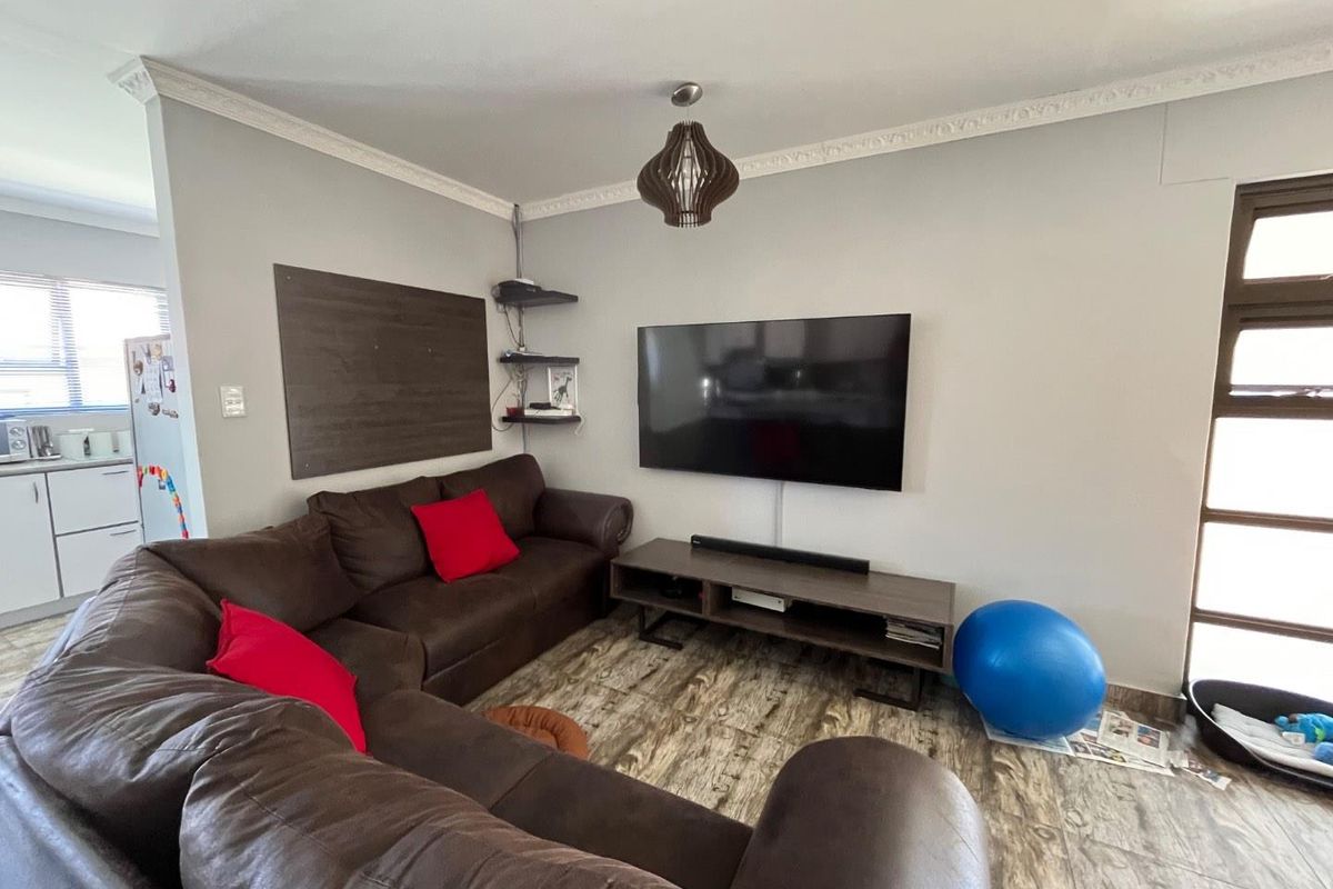 A comfortable living room featuring wood-look tile flooring, decorative crown molding, and a wall-mounted television setup, with an adjacent view into the kitchen area.