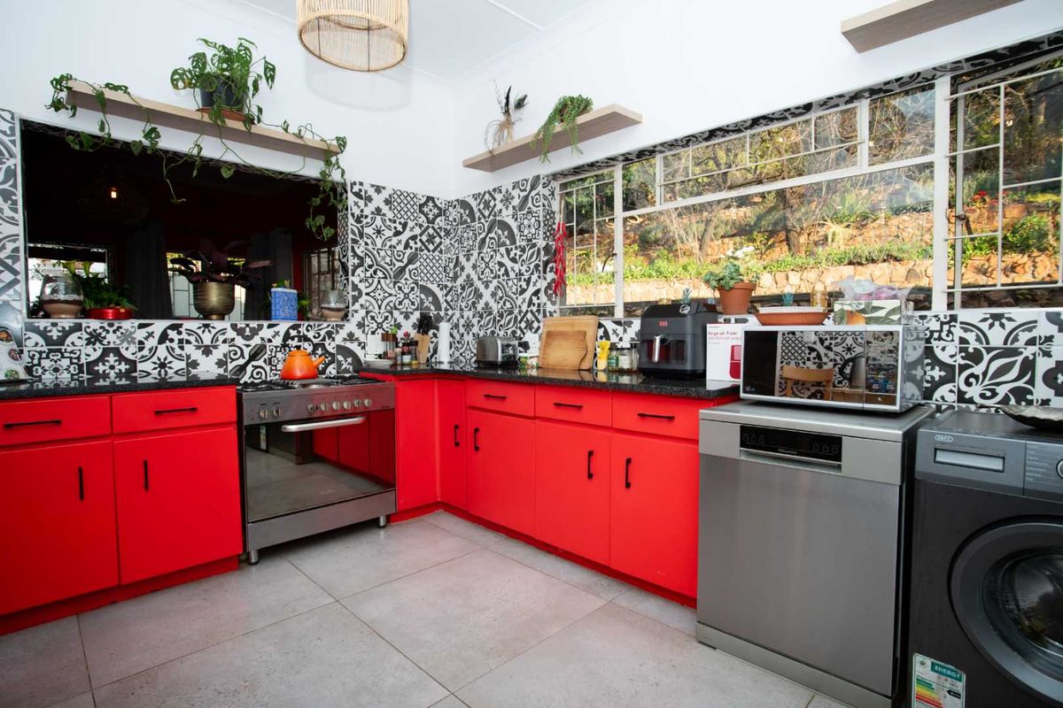 A vibrant kitchen featuring bold red cabinetry, contrasting patterned tile backsplash, granite countertops, and large windows providing natural light and garden views.