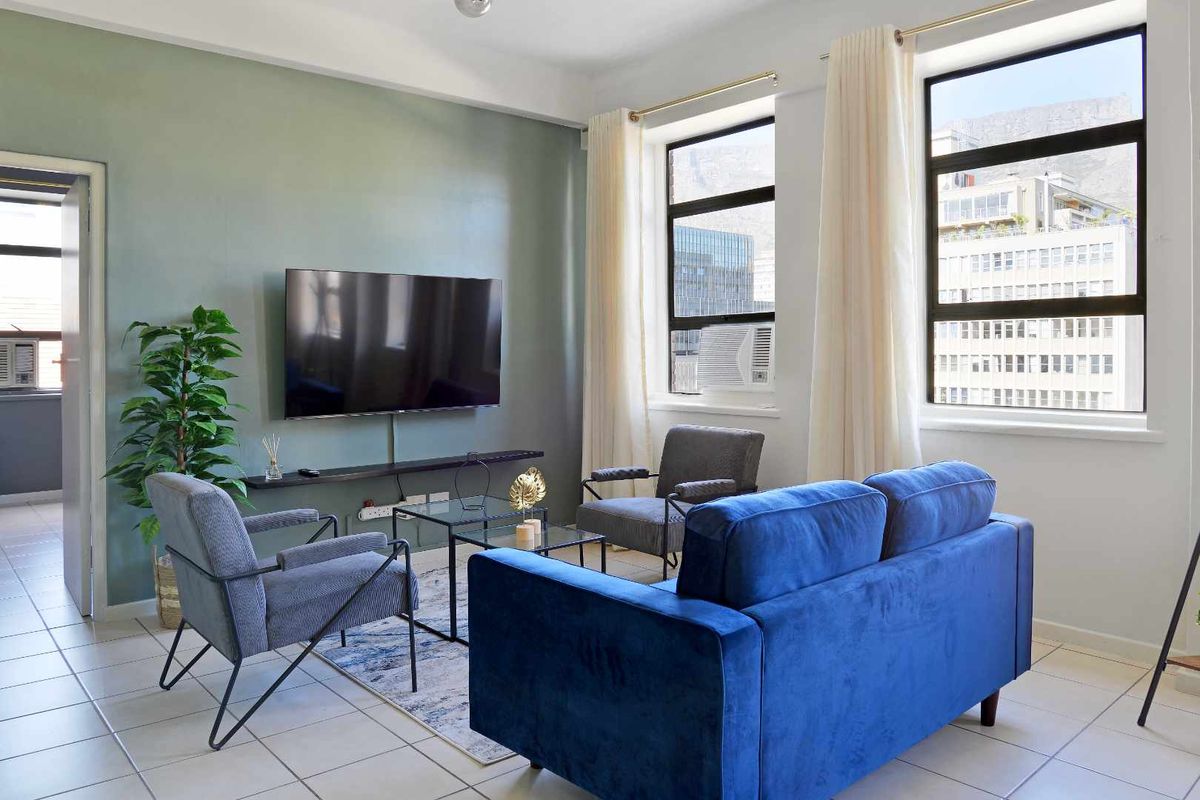 A bright living room featuring tiled flooring, large windows with natural light, and a wall-mounted television setup.