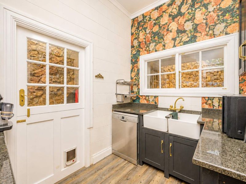 A well-appointed scullery featuring double farmhouse sinks, brass fixtures, granite countertops, and stylish floral wallpaper, providing a functional and aesthetic prep area.