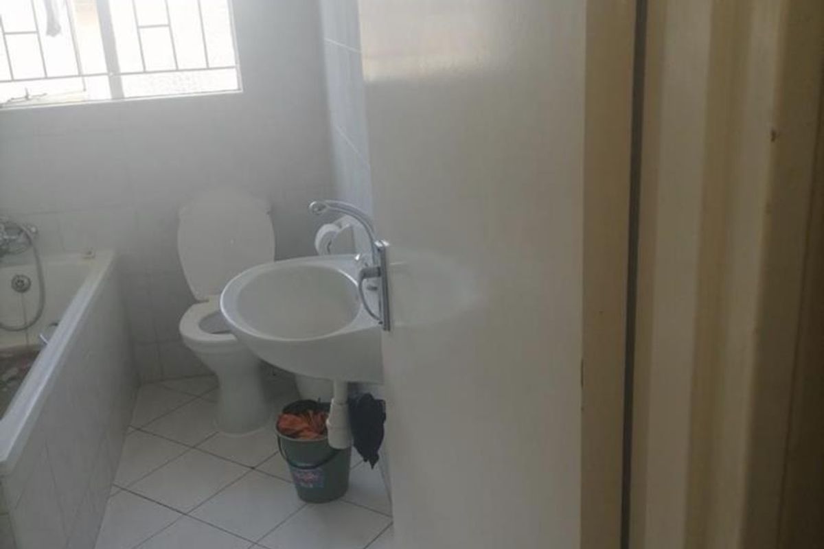 A functional bathroom featuring a bathtub, pedestal sink, and toilet, with white tiled flooring and natural light from a window with security bars.