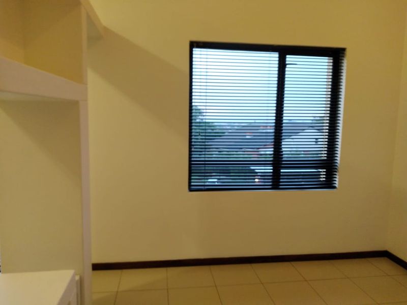 A bright, empty bedroom featuring neutral tiled flooring, built-in shelving, and a window fitted with dark venetian blinds.