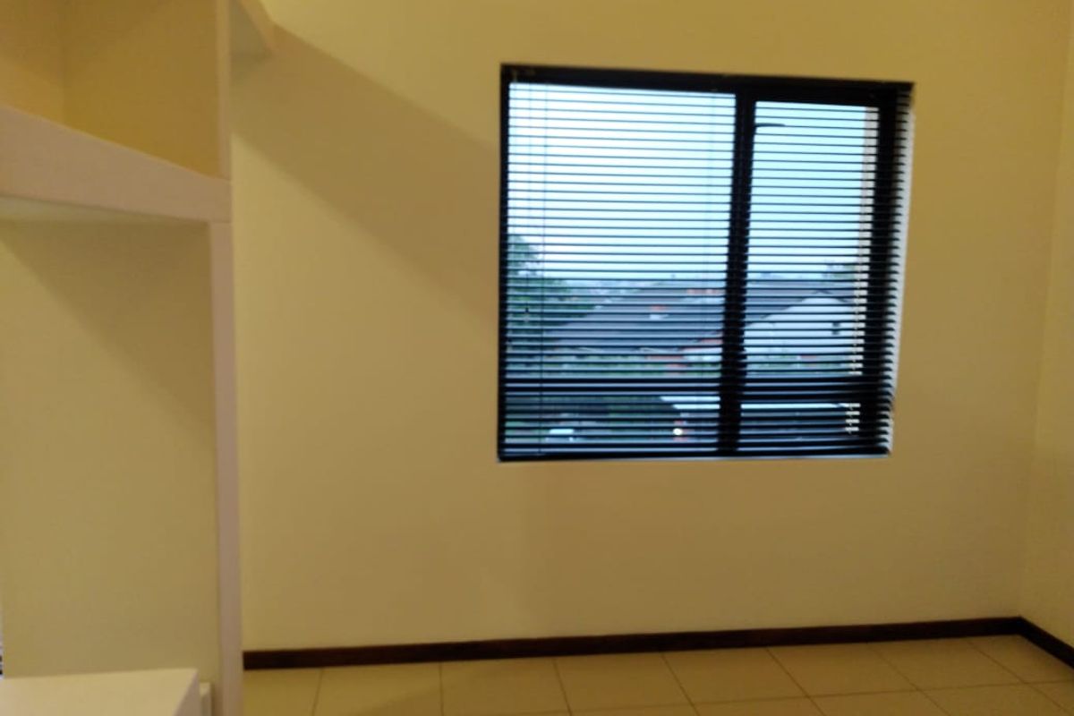 A bright, empty bedroom featuring neutral tiled flooring, built-in shelving, and a window fitted with dark venetian blinds.