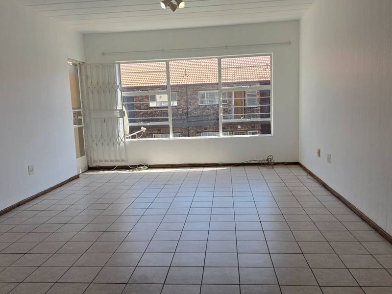 A spacious, empty living room featuring neutral tiled flooring, large windows for natural light, and a secure entrance gate.