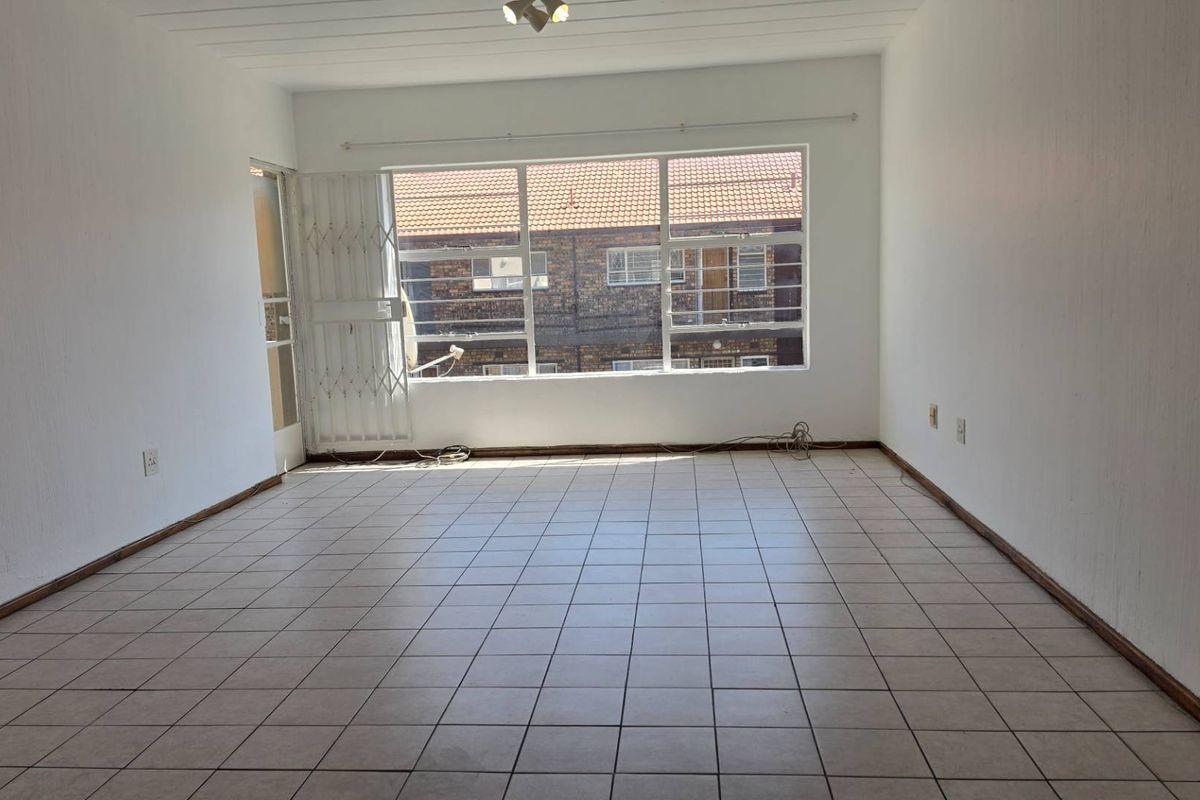 A spacious, empty living room featuring neutral tiled flooring, large windows for natural light, and a secure entrance gate.
