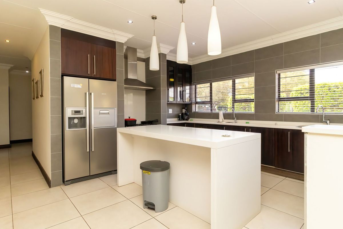 Modern kitchen featuring a central island, dark wood cabinetry, stainless steel extractor fan, double sink, and neutral tiled flooring with recessed and pendant lighting.