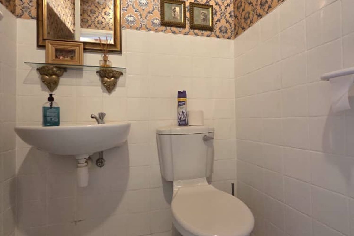 A compact bathroom featuring a wall-mounted basin, a standard toilet, patterned floor tiles, and decorative wallpaper above white wall tiling.
