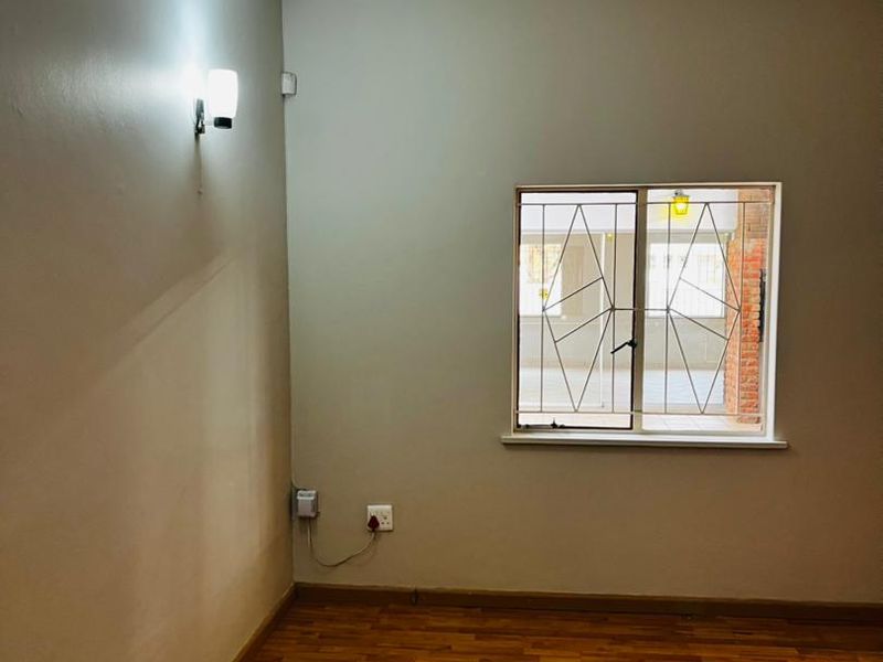 A clean, empty bedroom featuring warm-toned laminate flooring, a window with security bars, and a wall-mounted light fixture.