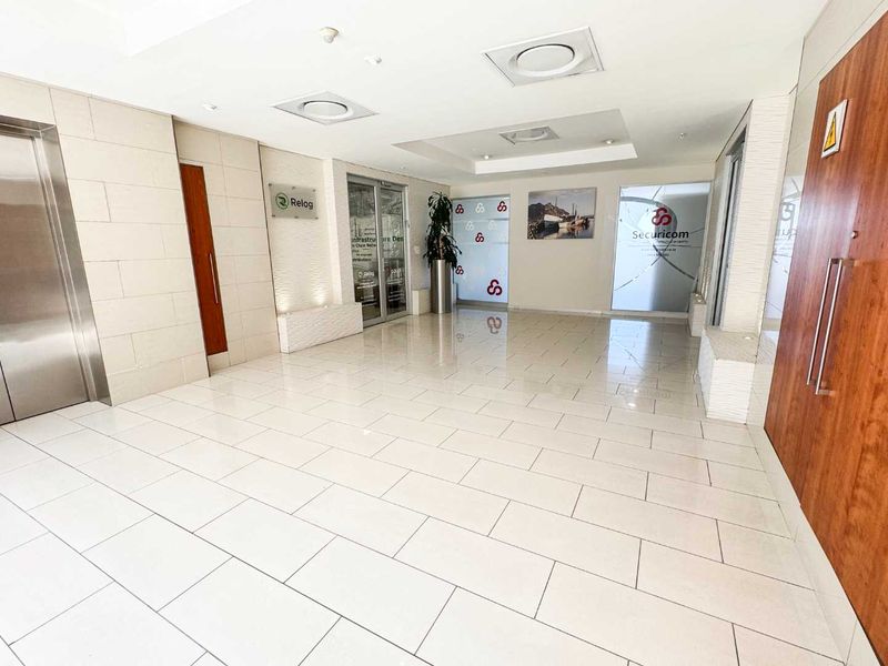 A bright and modern commercial building entrance lobby featuring tiled flooring, recessed ceiling lights, elevator access, and glass-fronted office suites.