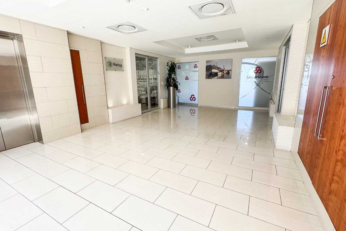 A bright and modern commercial building entrance lobby featuring tiled flooring, recessed ceiling lights, elevator access, and glass-fronted office suites.