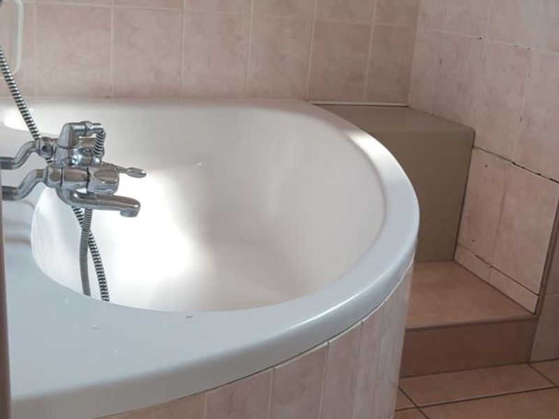 A bathroom featuring a built-in corner bathtub with a handheld shower mixer, finished with dated ceramic wall and floor tiling.
