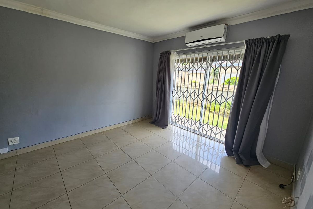 A bright living room featuring tiled flooring, a wall-mounted air conditioning unit, and sliding glass doors with security gates leading to the garden.