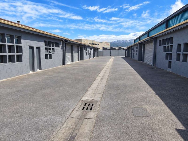 A well-maintained industrial complex exterior featuring asphalt driveway, concrete block walls, and multiple roller shutter access points.