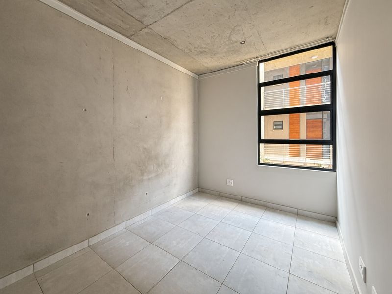 A modern, empty bedroom featuring an exposed concrete ceiling and feature wall, tiled flooring, and a large window providing natural light.
