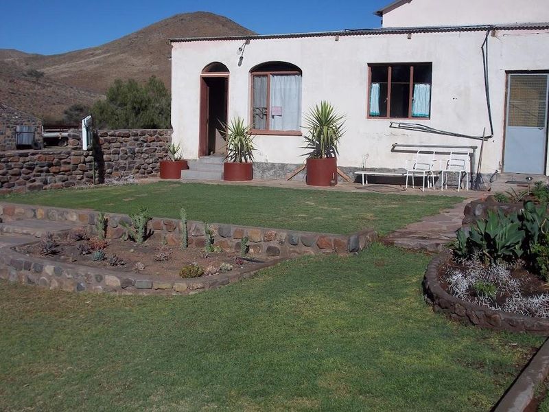 A rear view of a farm property featuring a landscaped garden with stone-walled flower beds, a lawn, and a simple exterior facade.