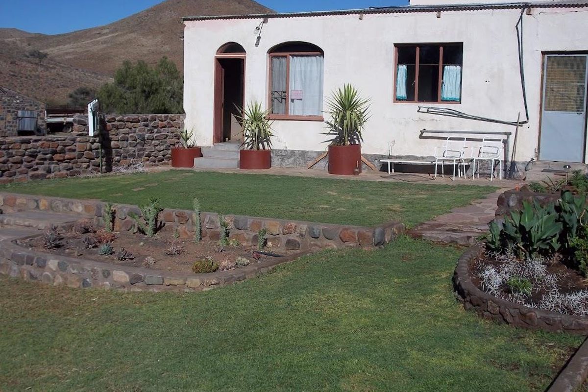 A rear view of a farm property featuring a landscaped garden with stone-walled flower beds, a lawn, and a simple exterior facade.