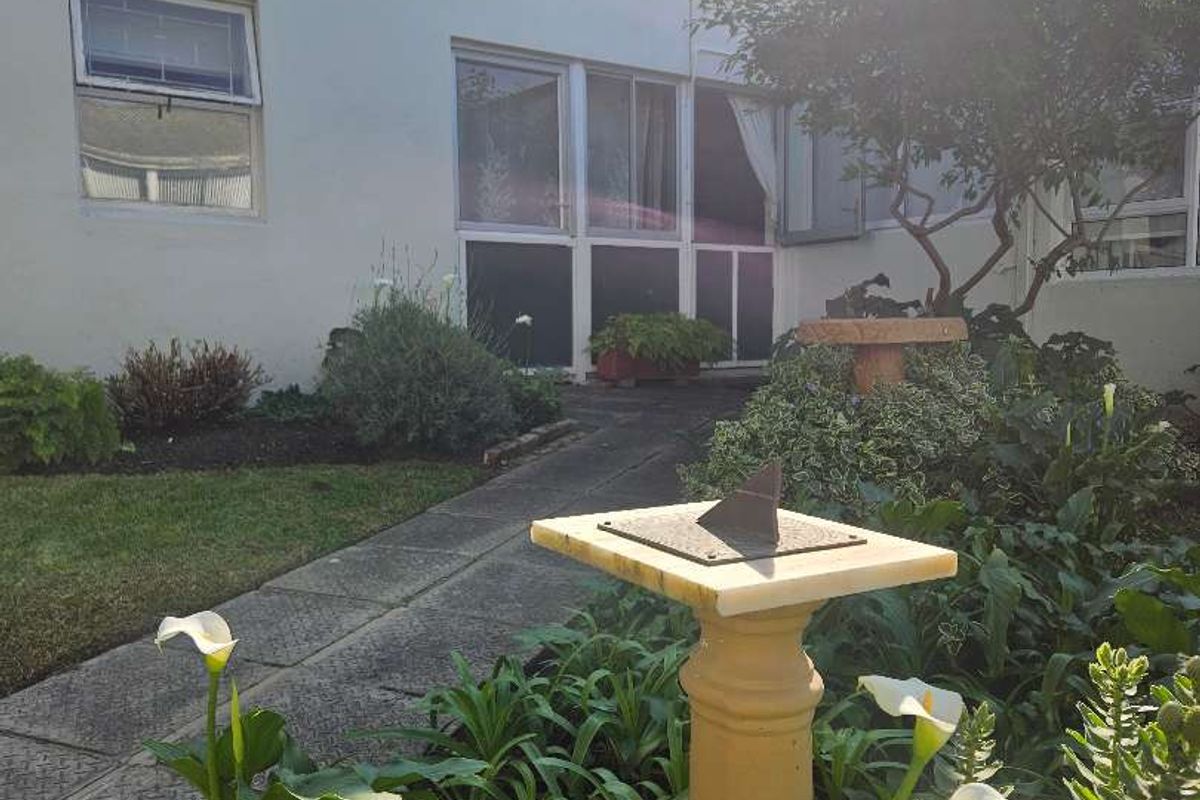A view of the rear exterior of the apartment building featuring a paved walkway, landscaped garden area, and sliding glass door access.