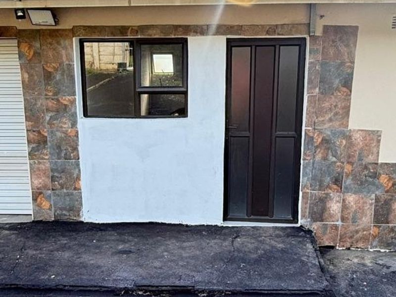 Exterior view of the apartment entrance featuring tiled wall accents, a security door, and an asphalt driveway area.