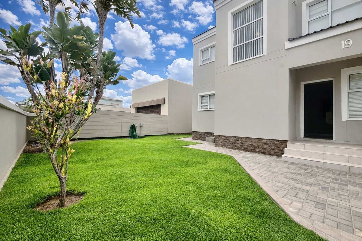 A well-maintained rear garden featuring a paved walkway, manicured lawn, and a modern two-story residential facade with stone cladding accents.