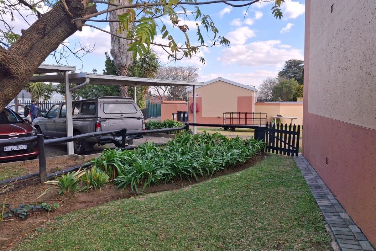 Exterior view of the townhouse property featuring a paved driveway, covered carport, landscaped garden bed, and a small wooden pedestrian gate.
