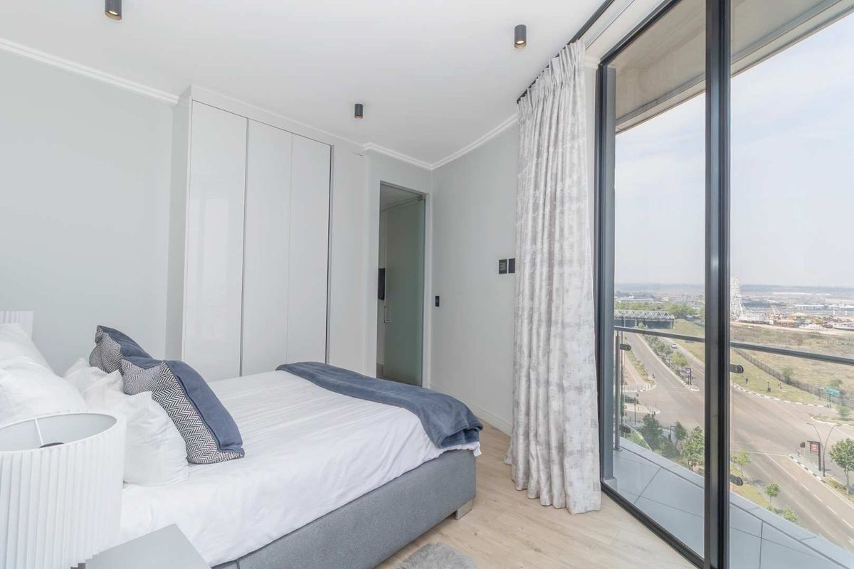 Modern bedroom featuring built-in wardrobes, laminate flooring, and floor-to-ceiling glass doors opening to a balcony with expansive city views.