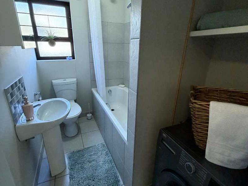 A functional bathroom featuring a pedestal basin, toilet, and a bathtub with shower attachment, complemented by tiled flooring and convenient built-in storage shelving.