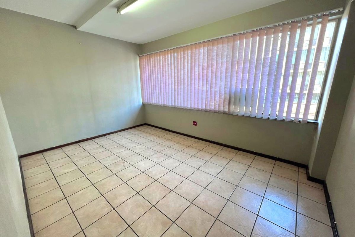 An empty office space featuring ceramic floor tiles, large windows with vertical blinds, and overhead fluorescent lighting.