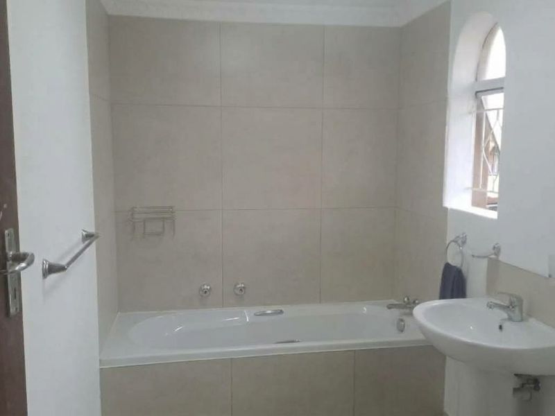 A functional bathroom featuring a built-in bathtub with tiled surround, a wall-mounted basin, and an arched window with security bars.