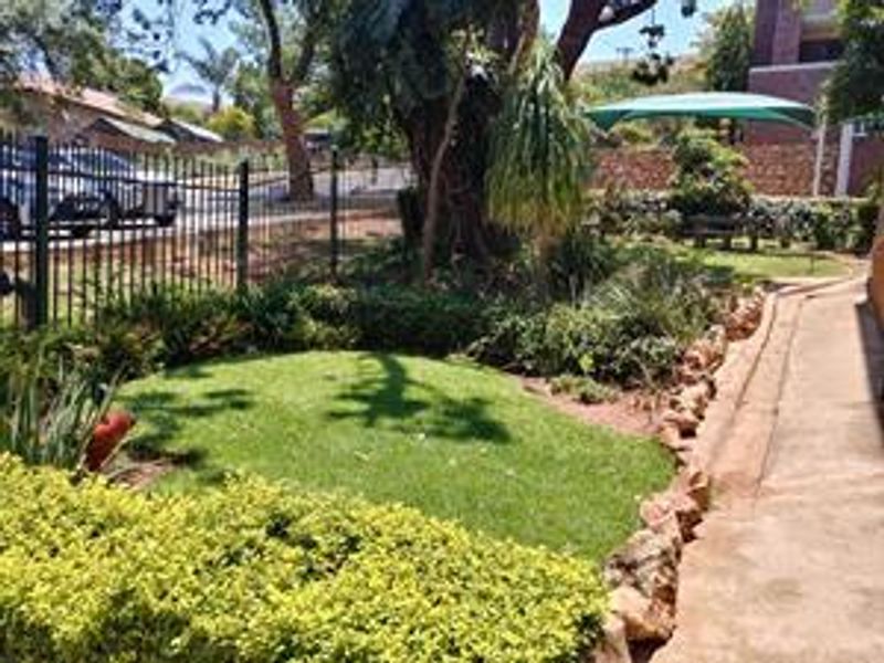 A well-maintained garden area featuring a concrete pathway, rock edging, and established trees, enclosed by metal perimeter fencing.