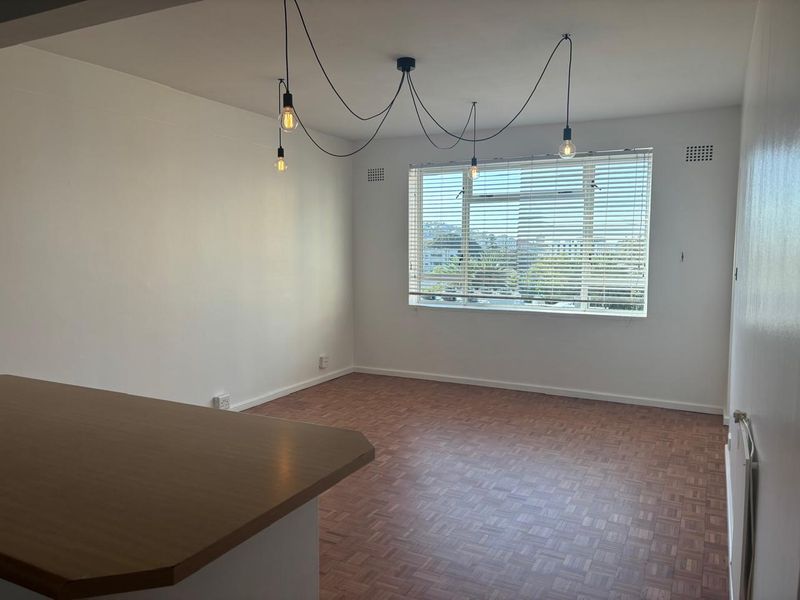 An open-plan living area featuring classic parquet flooring, a breakfast bar, and large windows with venetian blinds, offering a bright and versatile space.