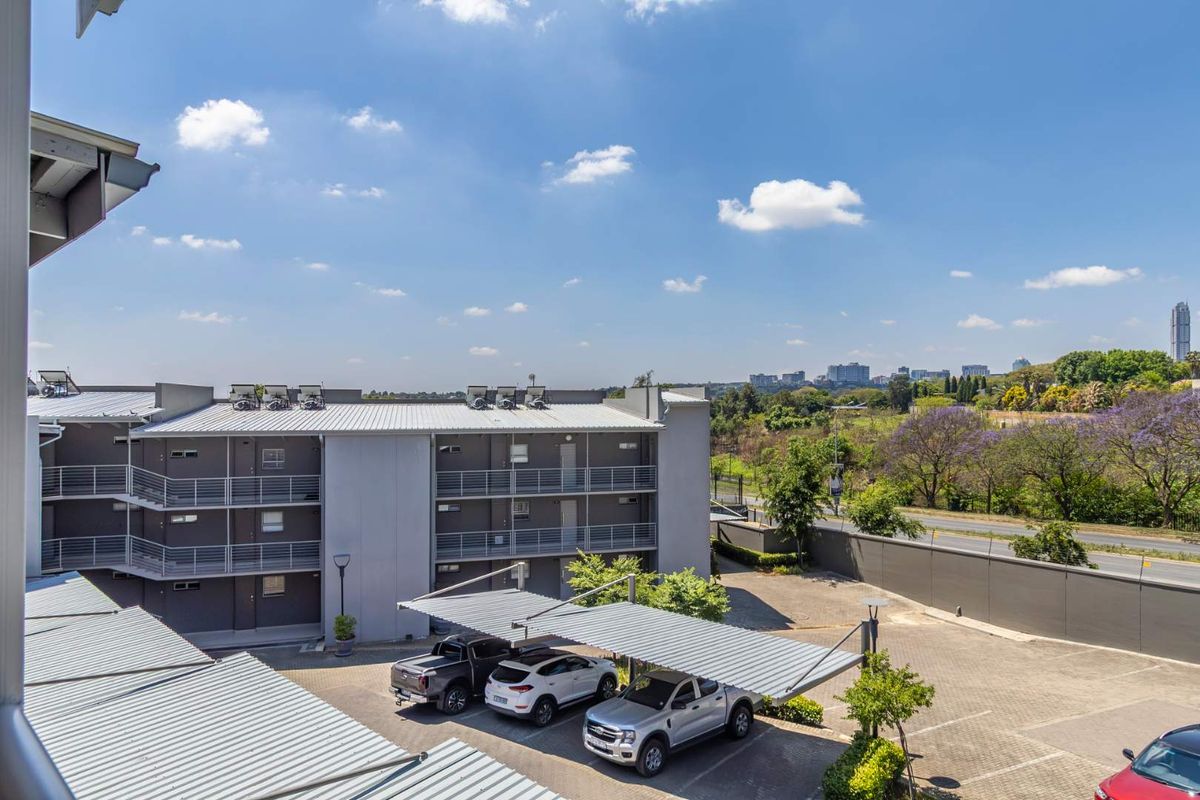 Elevated view of the apartment complex exterior, featuring covered parking bays, paved driveway, and surrounding landscaped grounds under a clear sky.