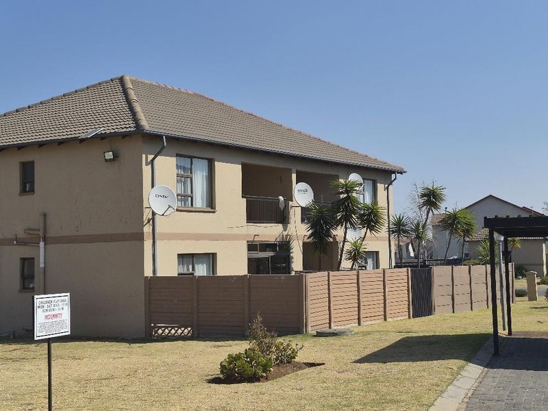 Exterior view of a multi-story apartment building featuring a tiled roof, private balconies, and perimeter fencing within a landscaped complex.