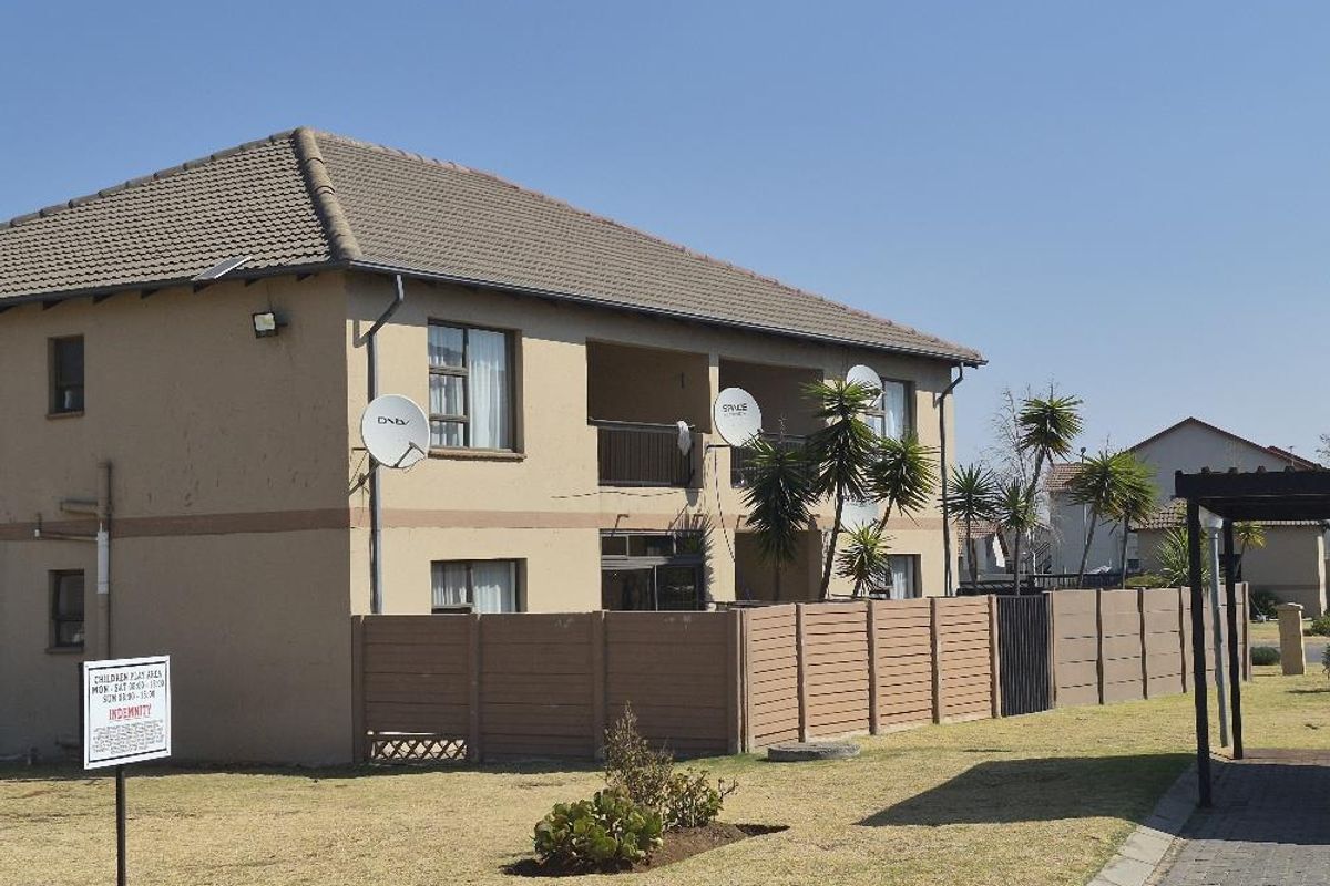 Exterior view of a multi-story apartment building featuring a tiled roof, private balconies, and perimeter fencing within a landscaped complex.