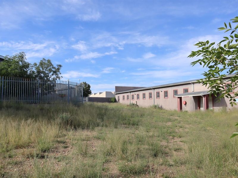 Rear exterior view of the property featuring a large unmaintained grass area, steel palisade fencing, and a long building structure with multiple windows.