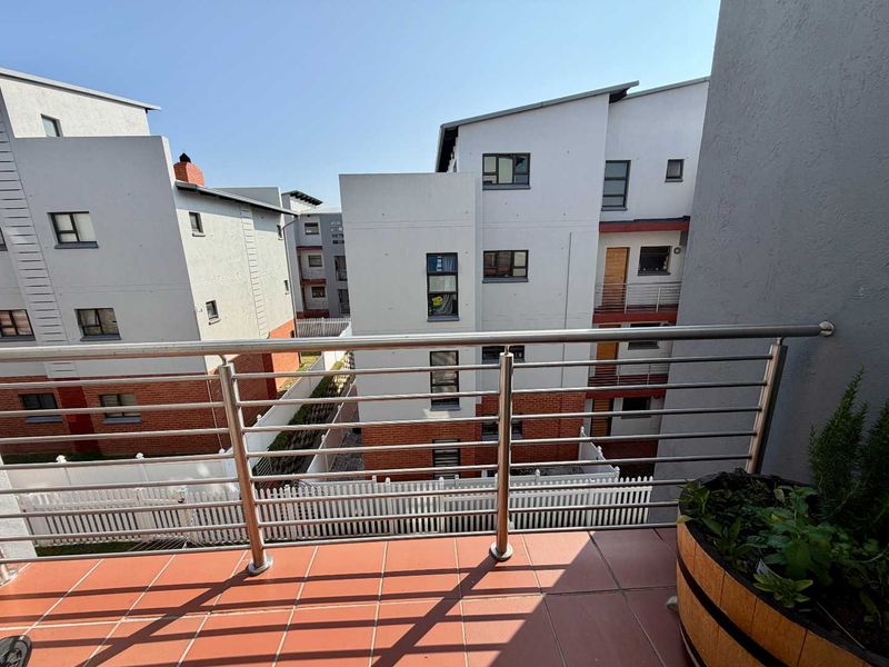 A private balcony featuring tiled flooring and a modern stainless steel balustrade, overlooking the apartment complex courtyard.