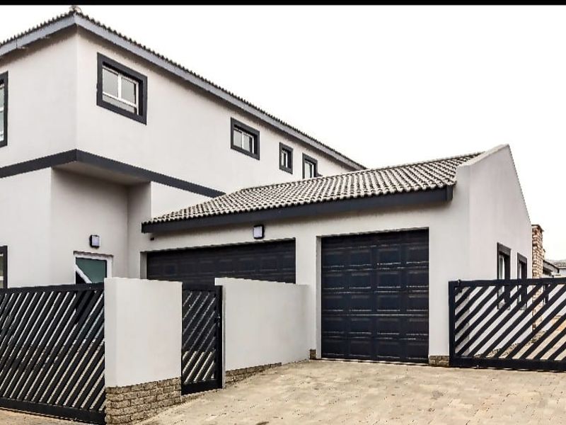 Modern double-story house featuring a double garage, paved driveway, and secure perimeter fencing with a contemporary grey and black exterior finish.