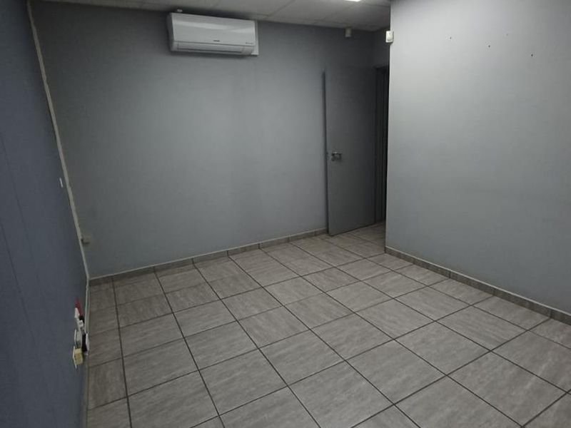 A clean, empty office space featuring grey walls, tiled flooring, a suspended ceiling with recessed lighting, and a wall-mounted air conditioning unit.