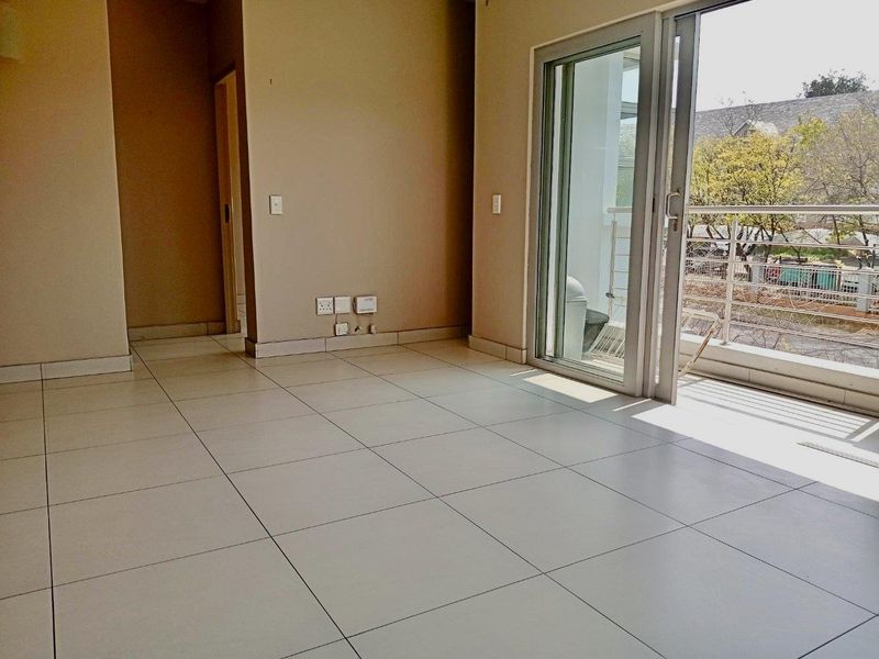 An empty, sunlit living area featuring neutral tiled flooring and a large sliding glass door providing direct access to an outdoor balcony.