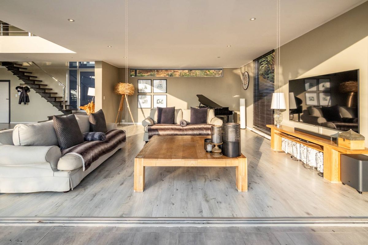 Spacious open-plan living area featuring modern timber flooring, recessed lighting, and a striking floating staircase, seamlessly connected to the outdoors via large sliding glass doors.