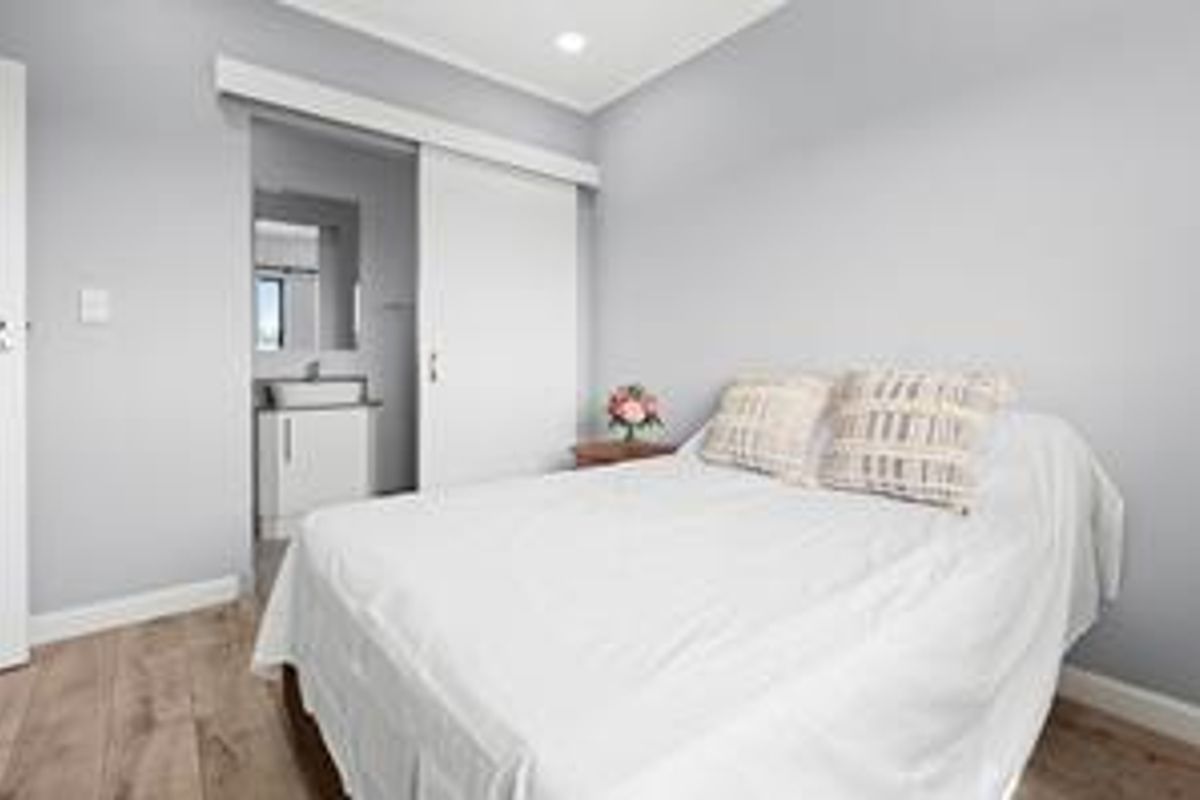 A modern bedroom featuring light grey walls, laminate flooring, recessed lighting, and a sliding door providing access to an en-suite bathroom.