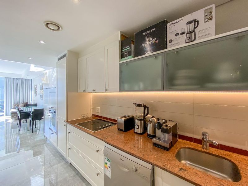 Modern kitchen featuring white cabinetry, stone countertops, integrated hob, and built-in oven, complemented by under-cabinet lighting and a tiled backsplash.