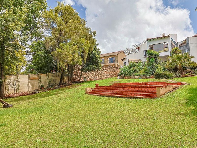 A spacious, sloped backyard garden featuring mature trees, a brick retaining wall, and terraced brick steps leading toward the multi-level residential structure.