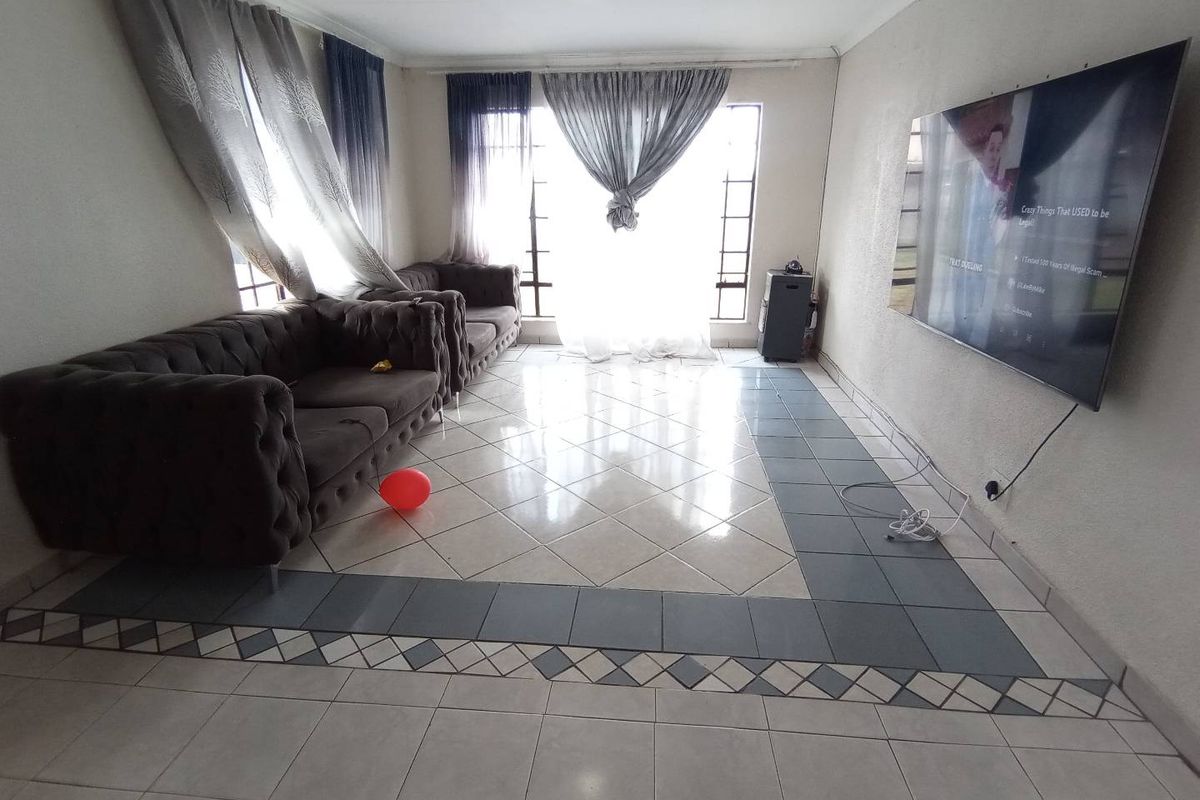 A spacious living room featuring large windows, tiled flooring with decorative border detailing, and wall-mounted television setup.