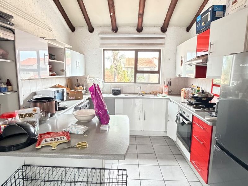 A functional kitchen featuring exposed timber ceiling beams, white and red cabinetry, tiled flooring, and a double basin sink with a view of the exterior.