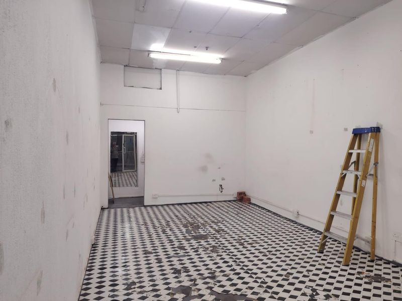 An empty retail space featuring checkered floor tiles, suspended ceiling with fluorescent lighting, and plain white walls requiring cosmetic attention.